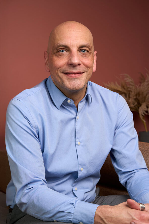 Portrait corporate d'un collaborateur chauve et souriant en chemise bleue sur fond terracotta, cabinet de conseil Paris